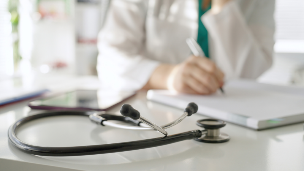 doctor writing patient notes in their office with a stethoscope on desk