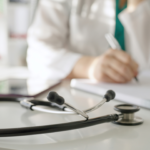 doctor writing patient notes in their office with a stethoscope on desk