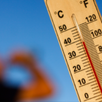 thermostat during heat wave showing temperature 100 derees as man drinks in background