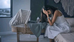 Woman with postpartum depression sitting with her head in her hand by baby bassinet crib